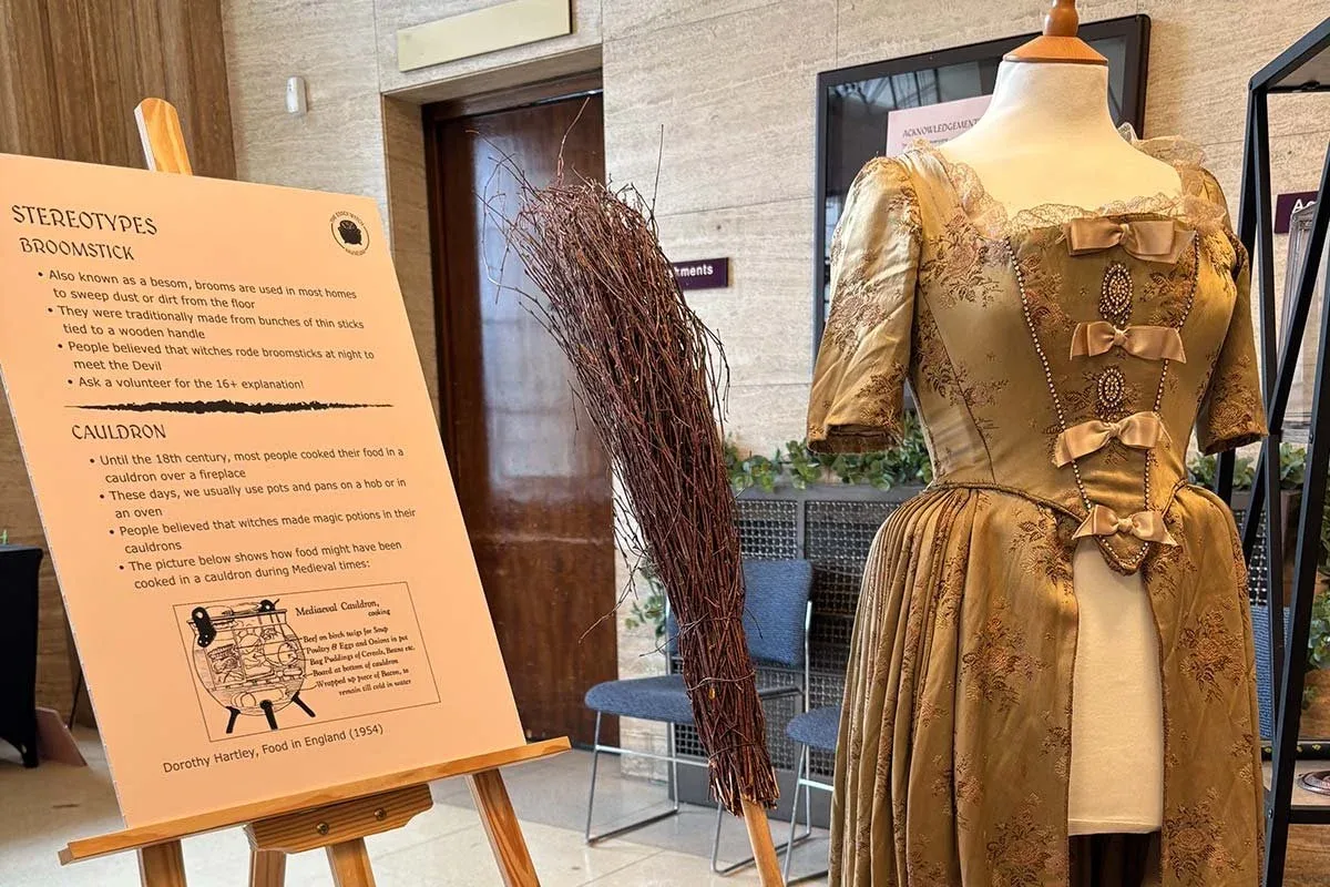 An Essex Witch Museum display in Shire Hall: a board on broomstick and cauldron stereotypes, a besom broom and a period gown