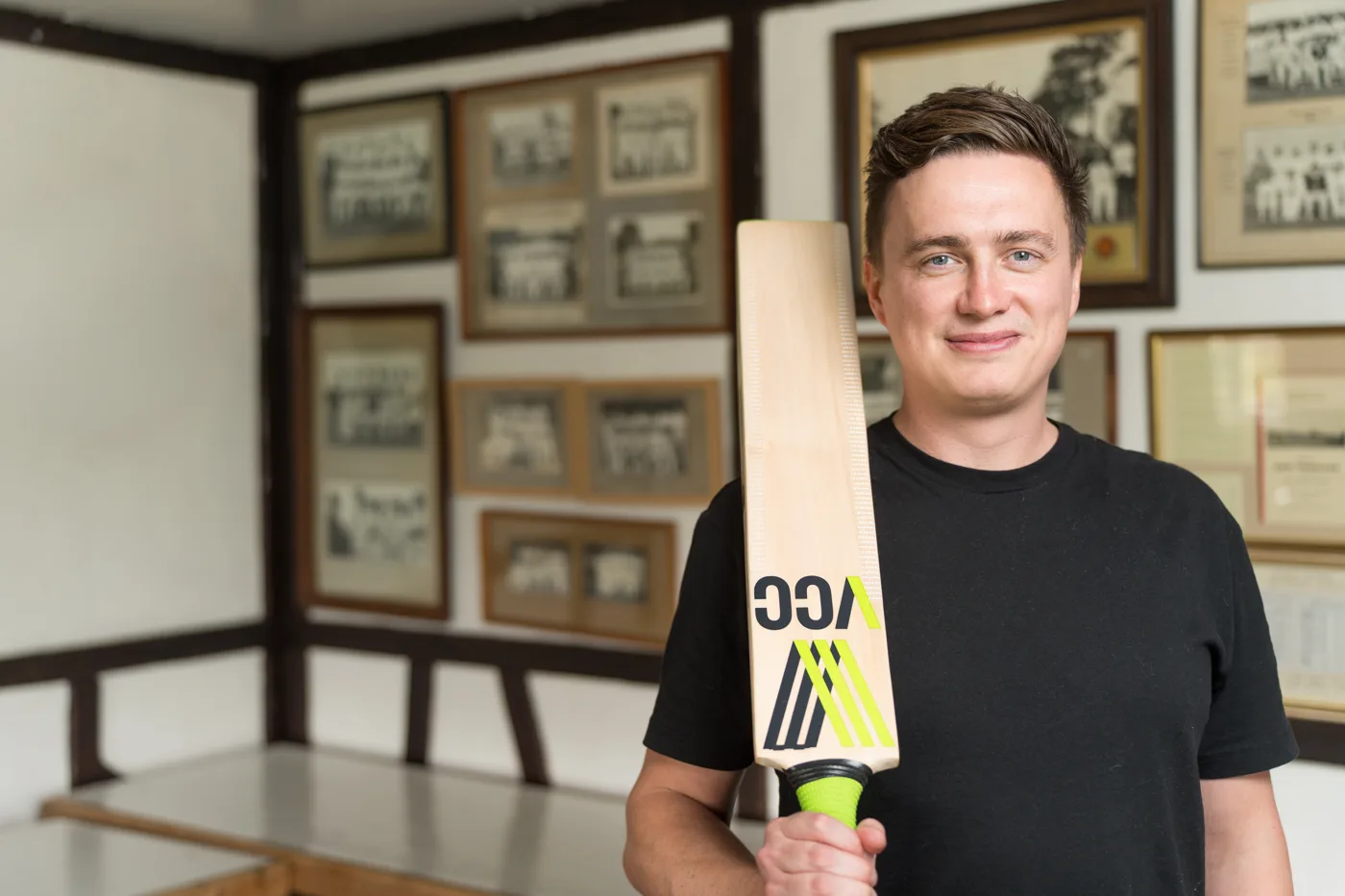 A man in a black T-shirt holding a Village Cricket Co. bat over his shoulder in a pavilion room hung with framed black and white team photographs