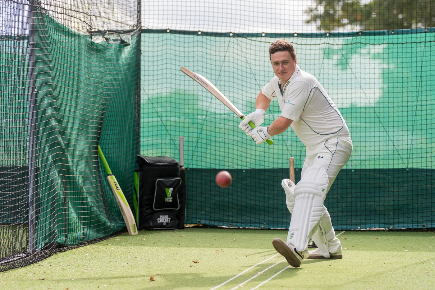 A batsman in whites and pads plays a shot in green-netted practice nets, with a Village Cricket Co. bat and kit bag propped against the netting behind him