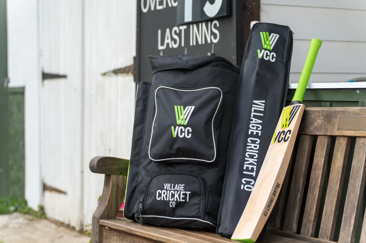 A black Village Cricket Co. kit bag, bat cover and bat with a lime green grip on a wooden bench in front of a scoreboard reading Last Inns