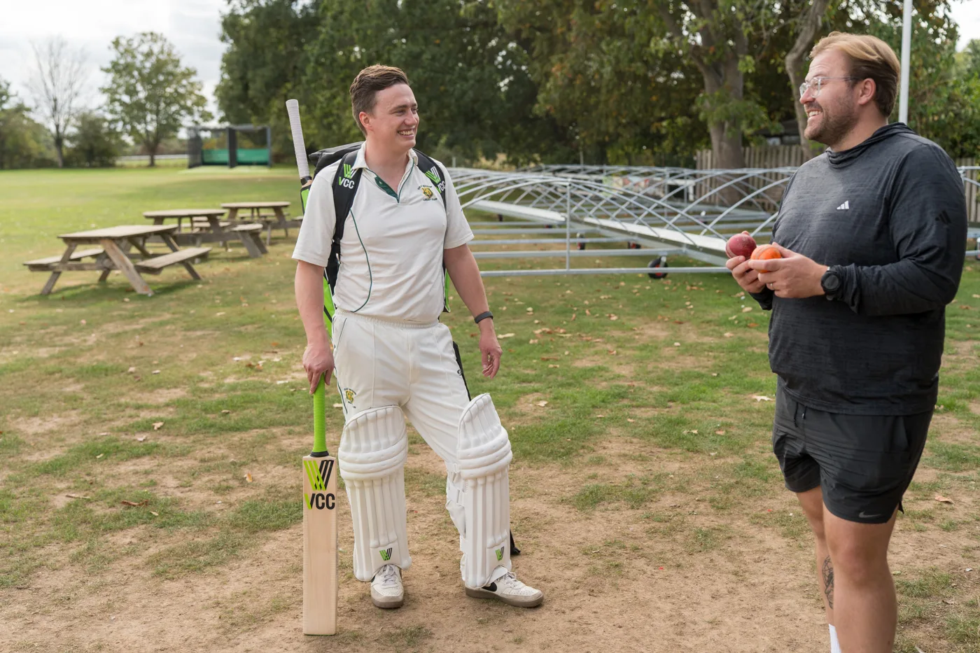Two cricketers talk on the outfield, one padded up with a Village Cricket Co. bat and kit bag