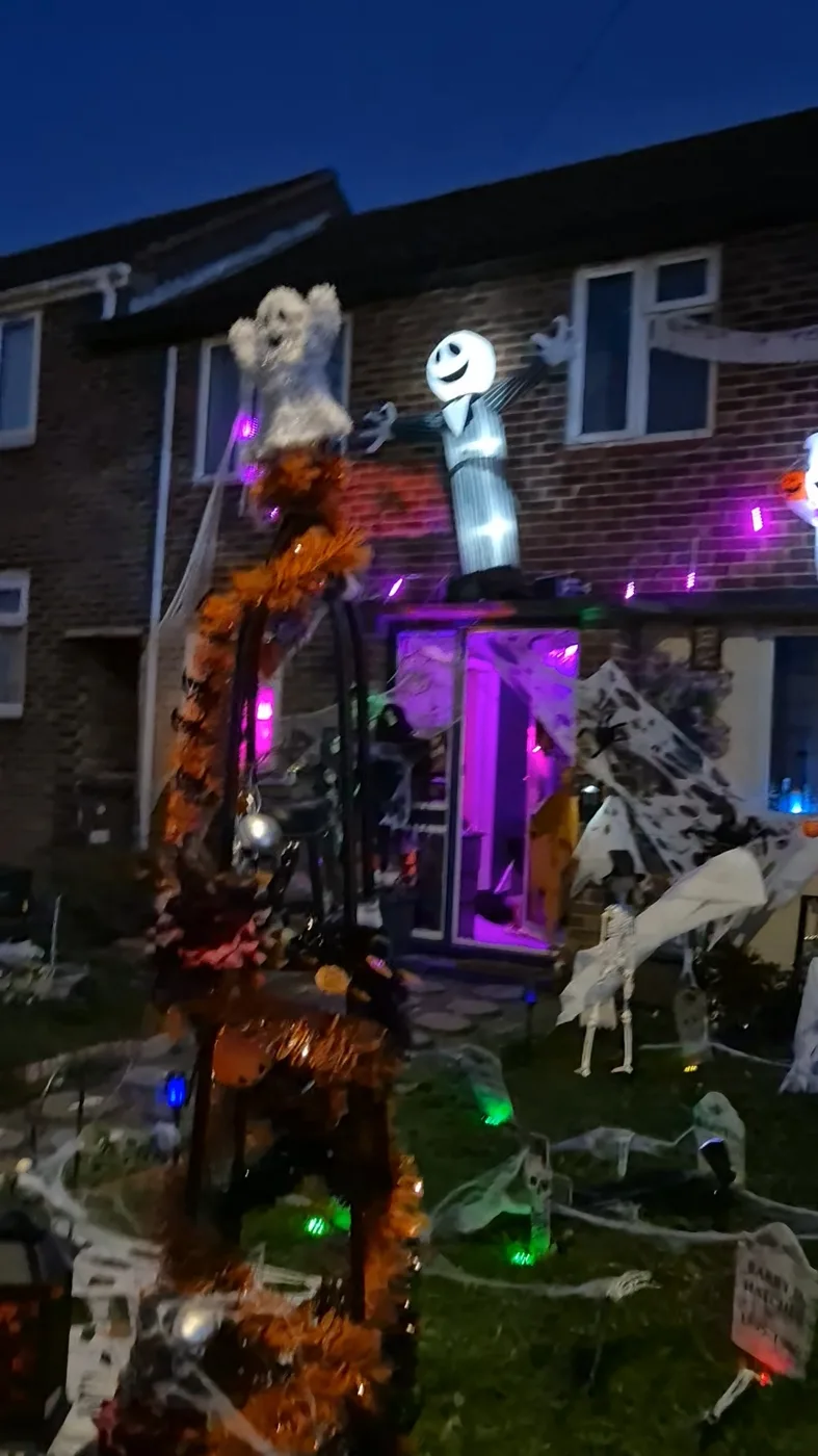 A tall frame wrapped in orange tinsel with a ghost on top, in front of a brick house hung with cobwebs and purple lights