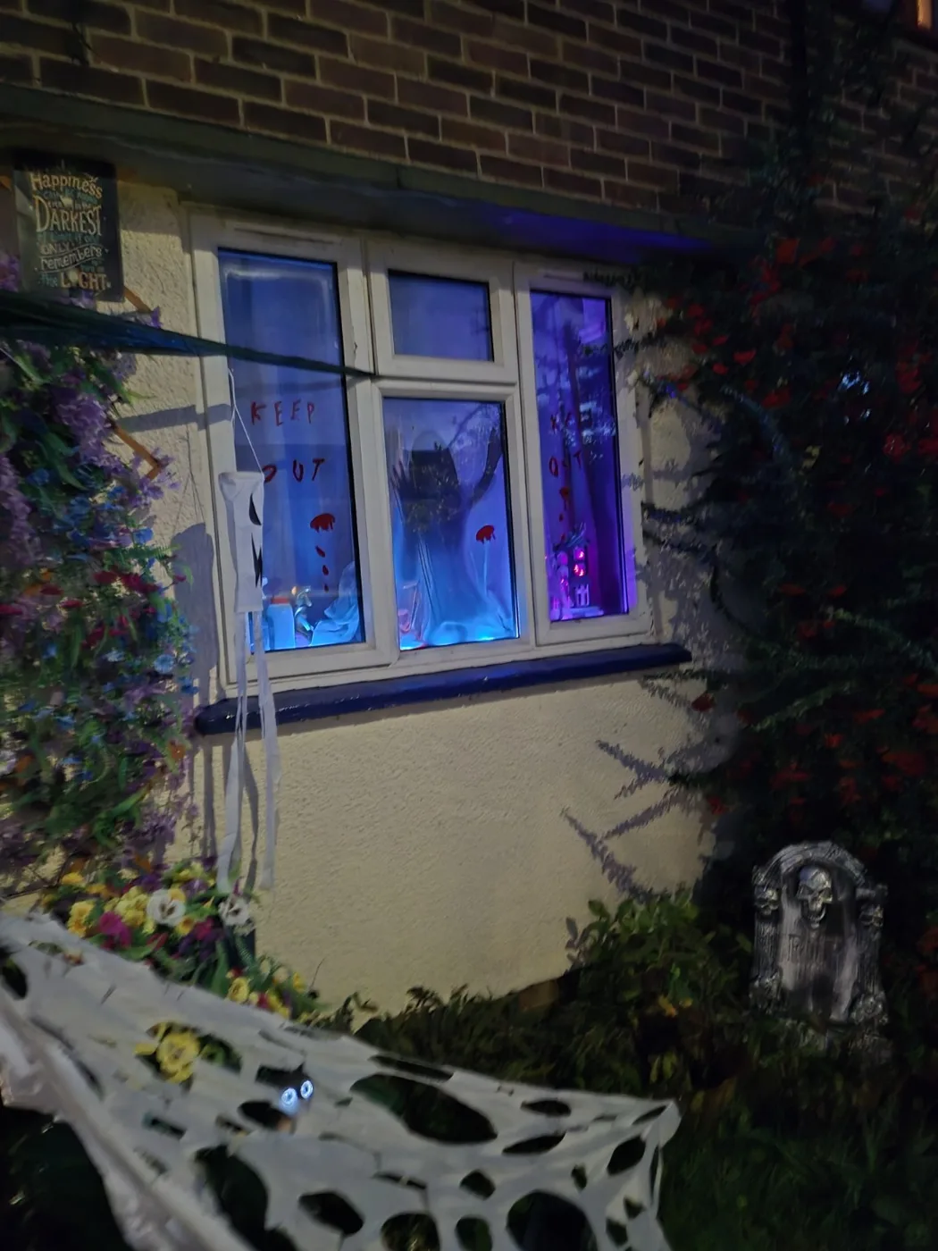 A figure lit in blue behind a ground floor window, with cobwebs and a gravestone in the flower bed below