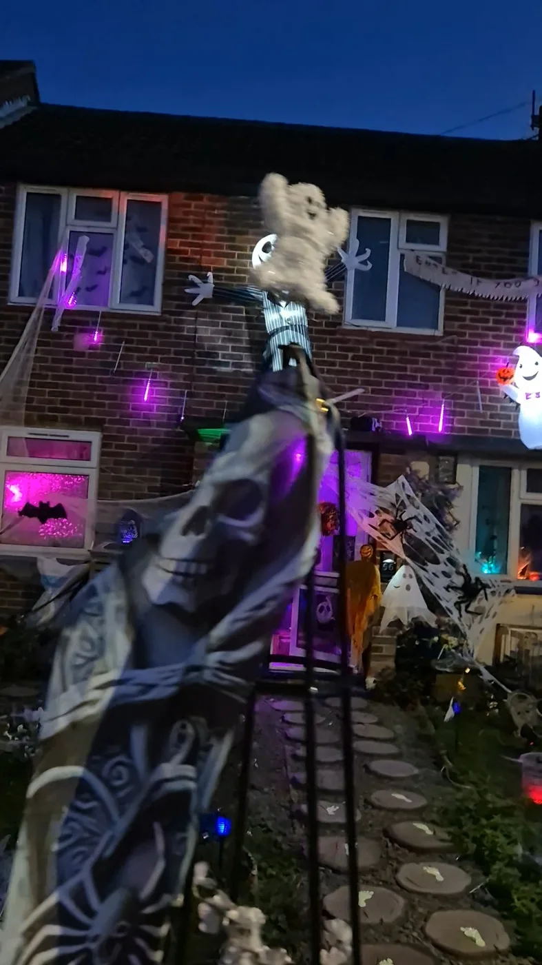 A skull-patterned cloth draped on a frame in the front garden, with a ghost on top and lit windows behind