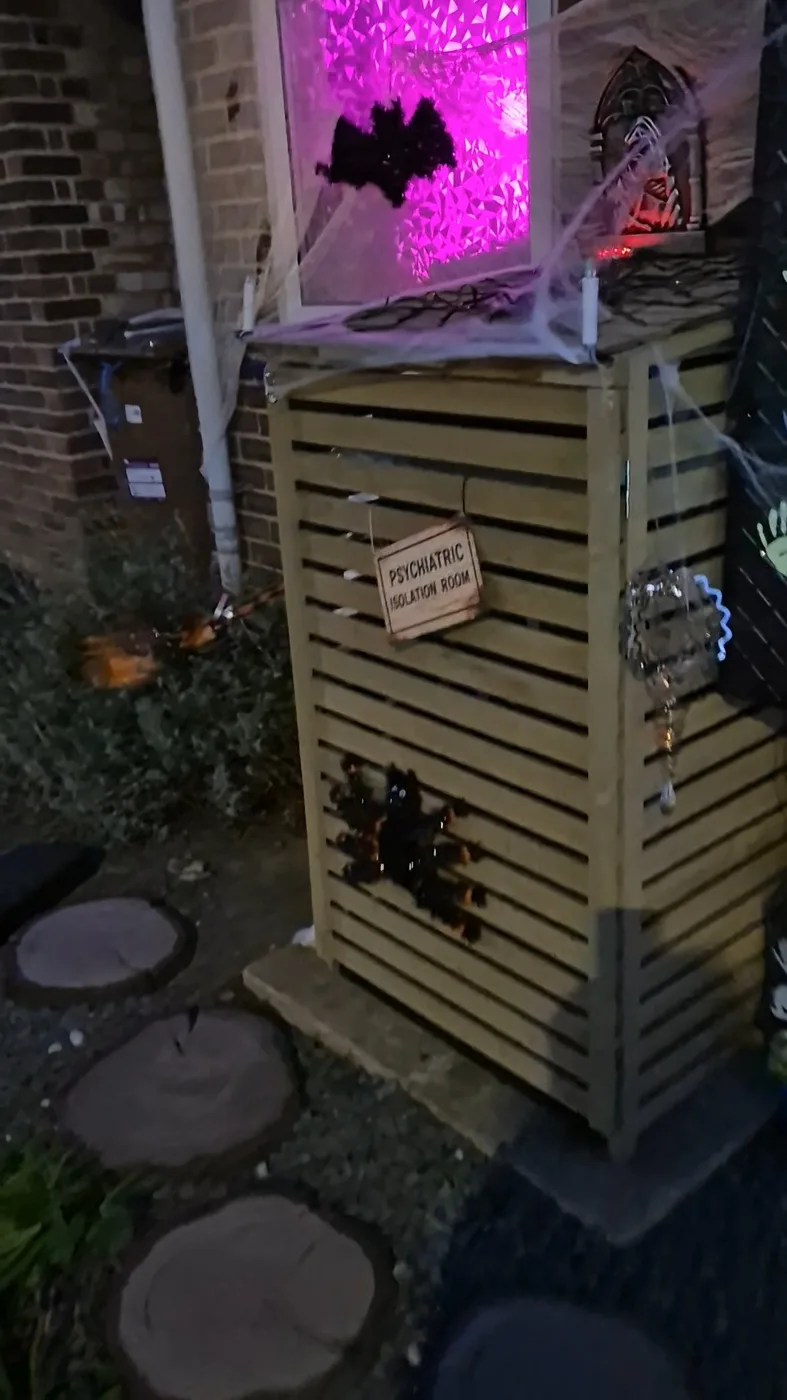 A wooden bin store covered in cobwebs with a sign reading Psychiatric Isolation Room and a spider on the door, under a pink lit window with a bat