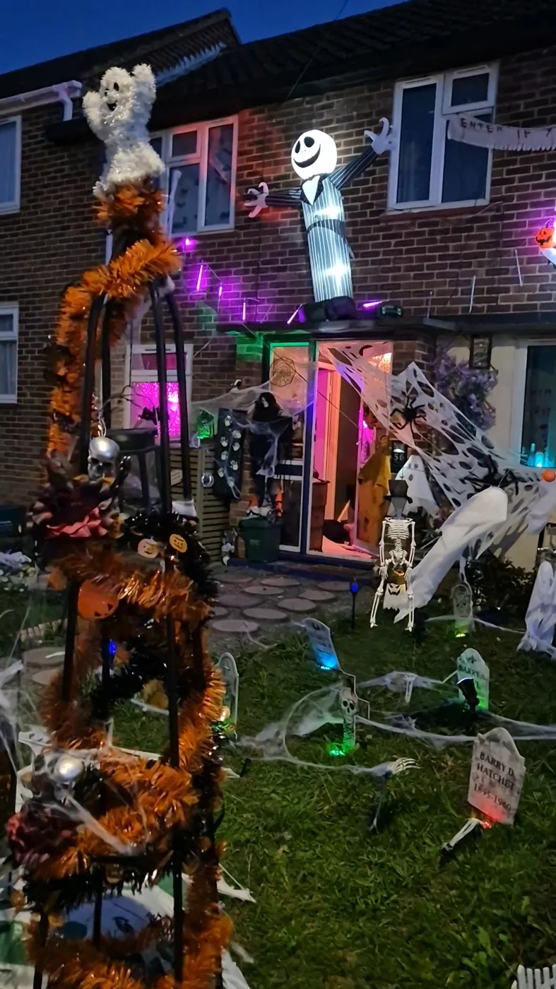 Wide view of the decorated front garden at dusk, with a tinsel frame, gravestones, coloured lights and ghosts on the brick wall
