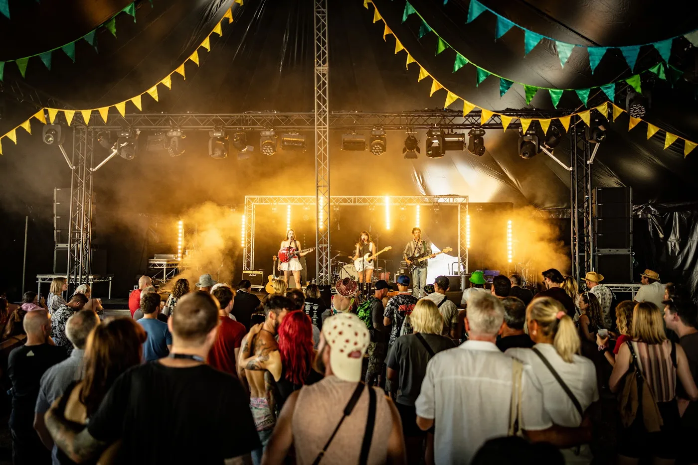 A band playing under lights in a tent stage hung with yellow and green bunting, the crowd in front