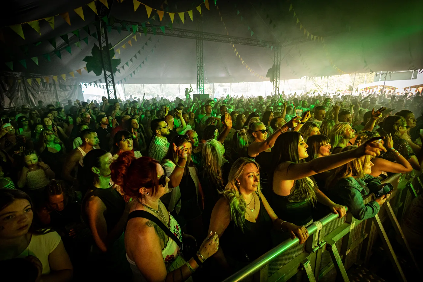 A packed crowd at the barrier inside a tent stage under green lights
