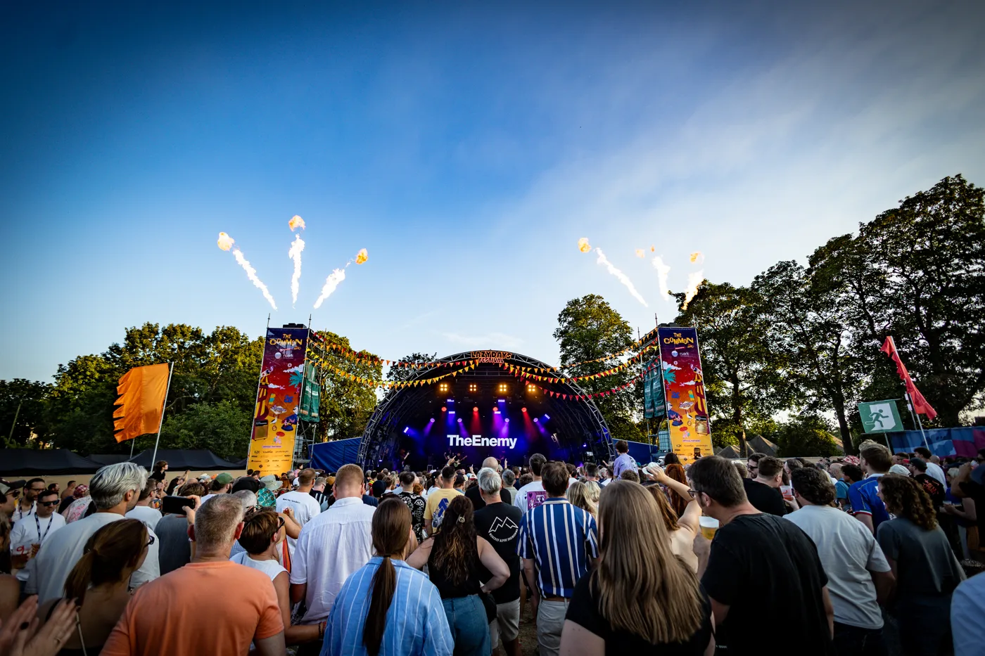 The main stage in daylight with pyrotechnics firing above it and a large crowd in front
