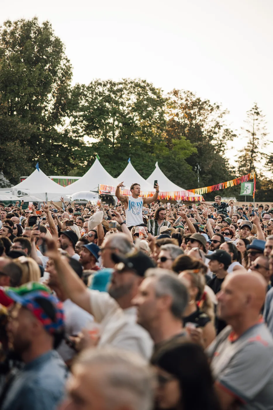 A dense daytime crowd in front of white marquees and trees