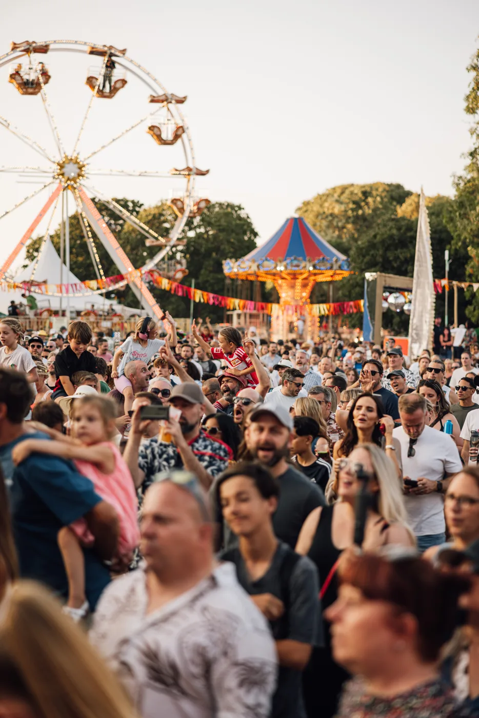 A crowd of festival-goers with a big wheel and a fairground ride behind them