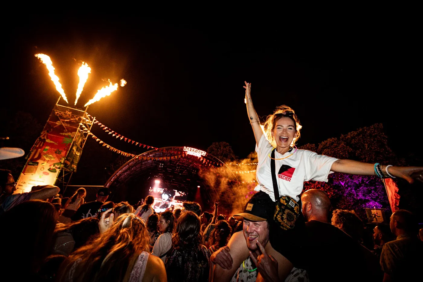 A woman on someone's shoulders with her arms raised in a crowd at night, flame jets firing beside the main stage