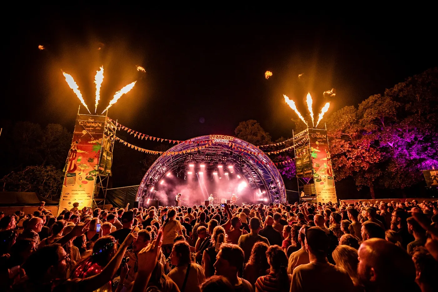 A packed crowd facing a lit main stage at night with flame towers on either side
