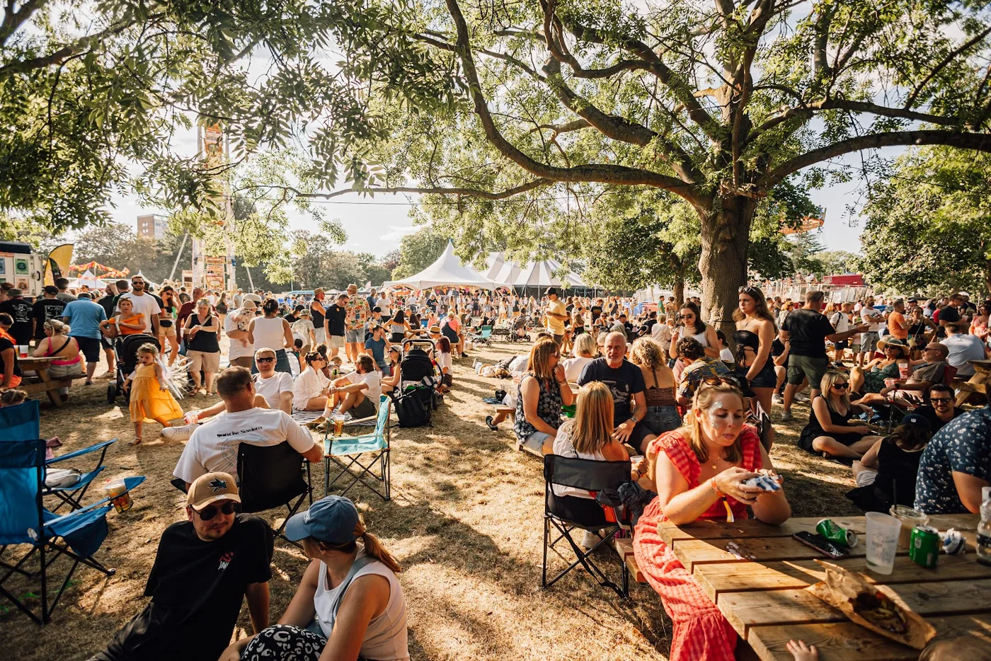 Festival-goers sitting at picnic tables and on camping chairs in the shade of large trees