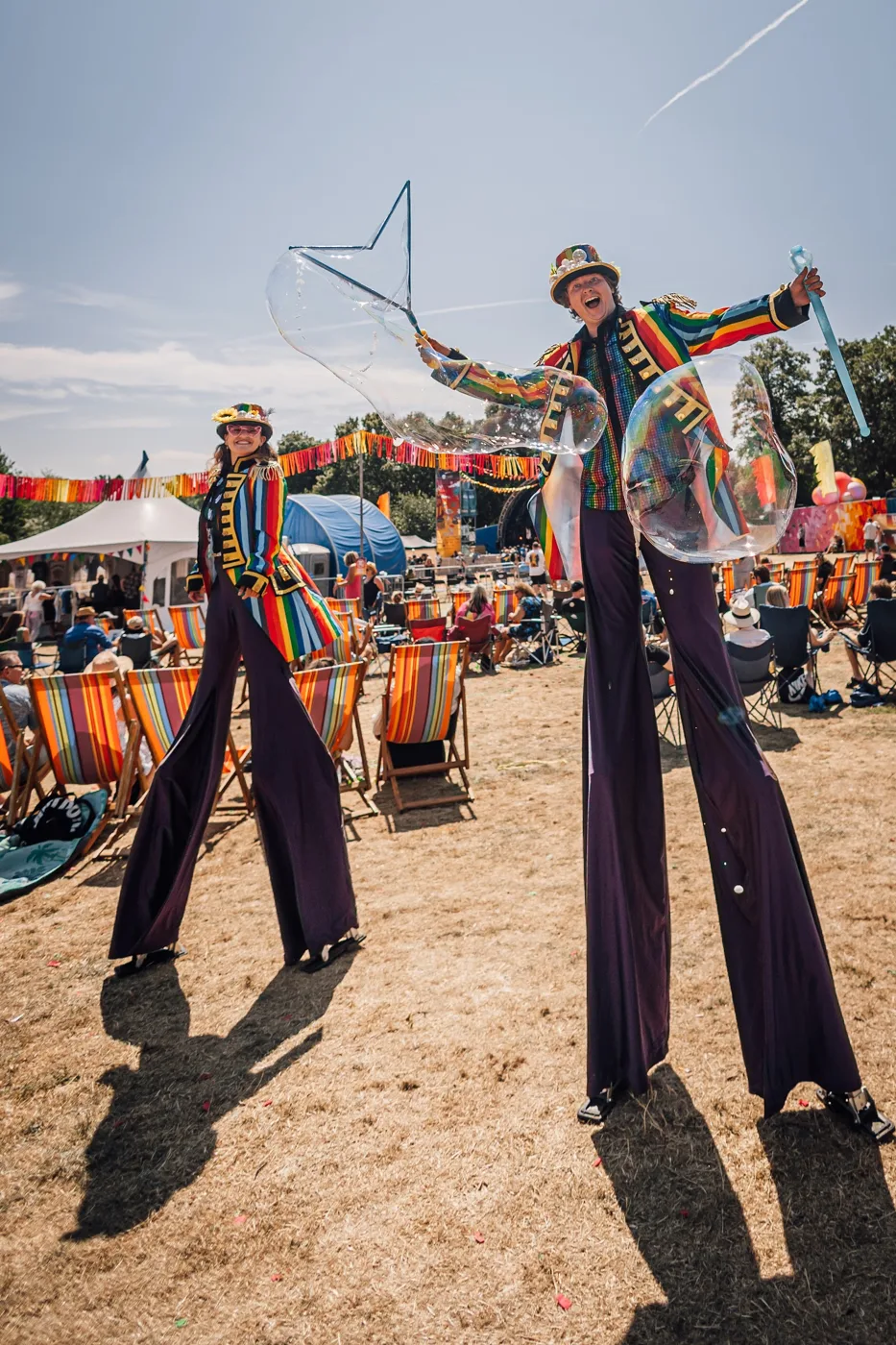 Two stilt walkers in rainbow-striped jackets and top hats, one blowing giant bubbles
