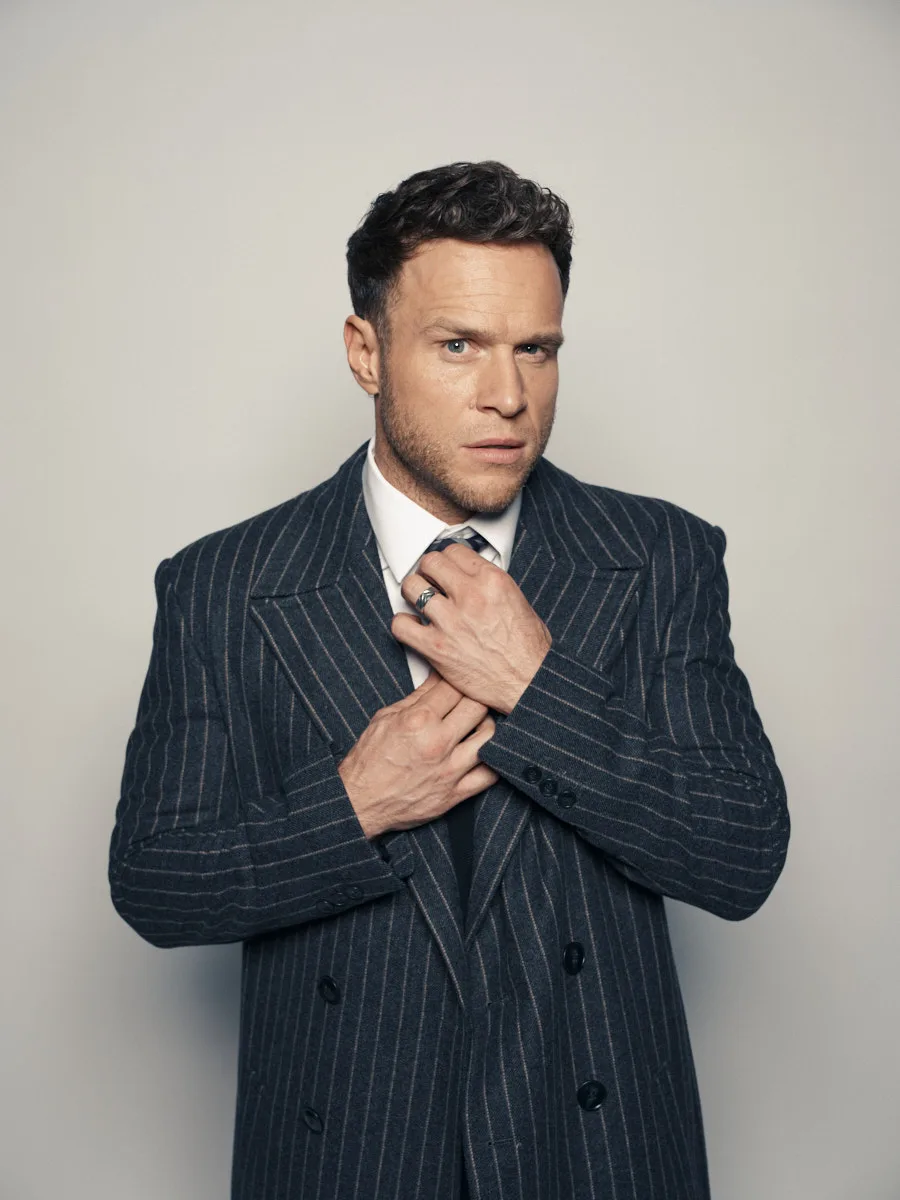 Olly Murs in a dark pinstripe double-breasted suit, adjusting his tie, against a pale grey wall