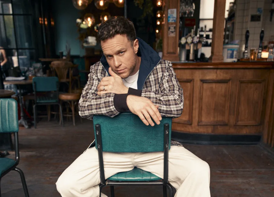 Olly Murs leaning on the back of a green chair inside a pub, with the bar behind him