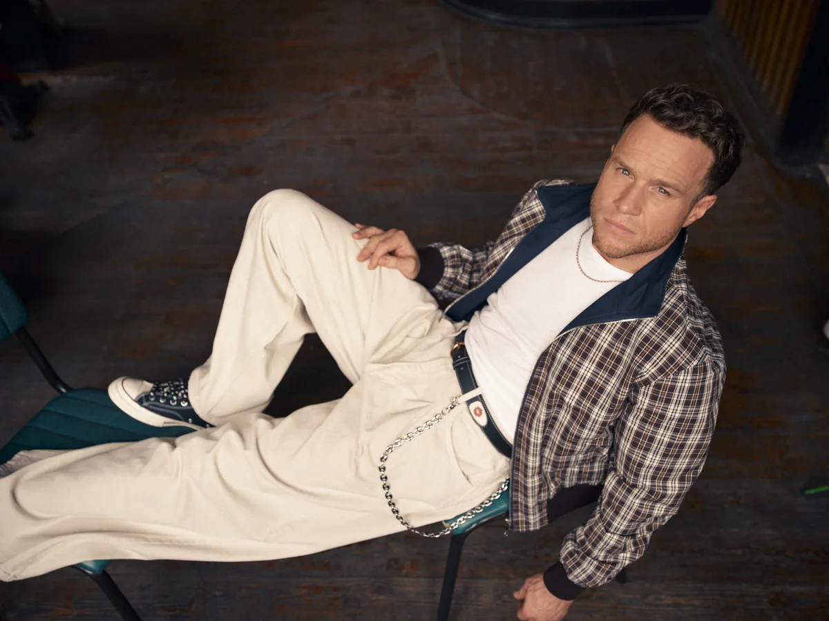 Olly Murs reclining across a chair on a wooden floor, in a check jacket and cream trousers